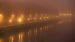 Streetlights glow through dense fog by the water.