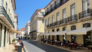 A flat, wide, pedestrianised street in Lisbon's Baixa district, with classic Pombaline architecture and outdoor cafes, under a clear blue sky.