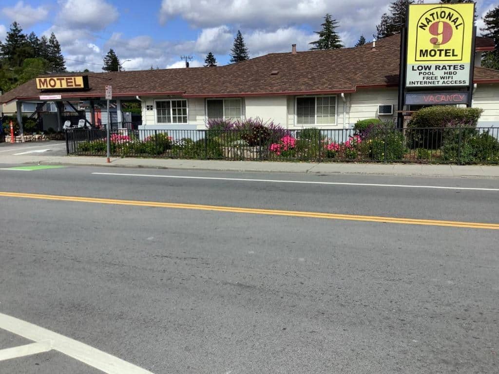 Daytime exterior of National 9 Motel in Santa Cruz showing the iconic yellow-and-red roadside sign, flowering garden and shingled roofline behind a low picket fence on Mission Street