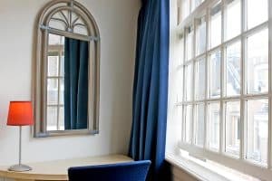 Room corner with an arched rattan mirror and a red lamp beside a window onto the brick buildings opposite, Hotel De Koopermoolen, Amsterdam