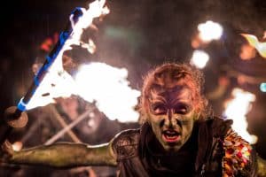 A Samhuinn Fire Festival performer in green face paint holding a lit fire staff in the rain at night