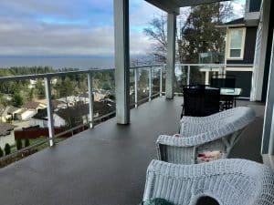 The shared balcony at Ocean Melody Vacation Rooms, wicker chairs behind a glass balustrade above treetops and the Salish Sea