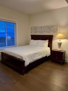 A guest room with a bed, a bedside lamp and a bright window over a hardwood floor