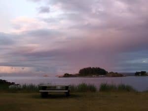 The shoreline at Neck Point Park in Nanaimo, a picnic bench on the grassy foreshore looking across calm water to a wooded island under a pink evening sky