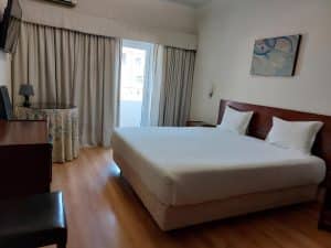 A guest room at the Petrus Hotel in Chaves with a wide bed, wooden floor and full-length curtains at the balcony window