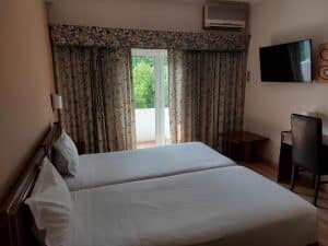 A twin room at the Petrus Hotel with two beds, patterned curtains, a wall-mounted television and doors onto a balcony