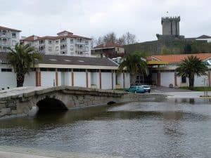 The thermal baths building at the Termas de Chaves beside the river, with palm trees along the front and the castle keep on the hill behind