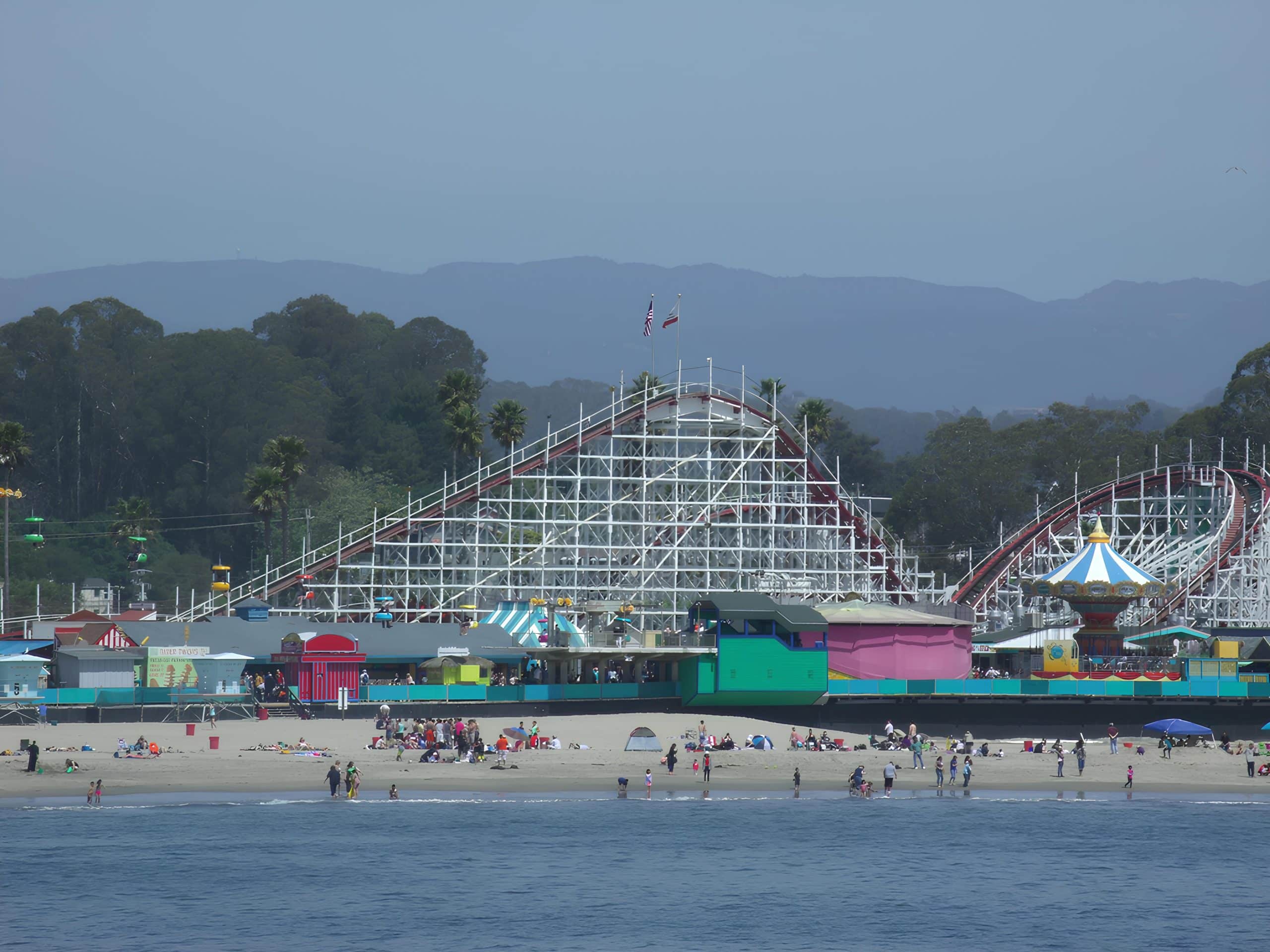 The white wooden Giant Dipper coaster rising behind the painted arcades of Santa Cruz Beach Boardwalk, seen from the water