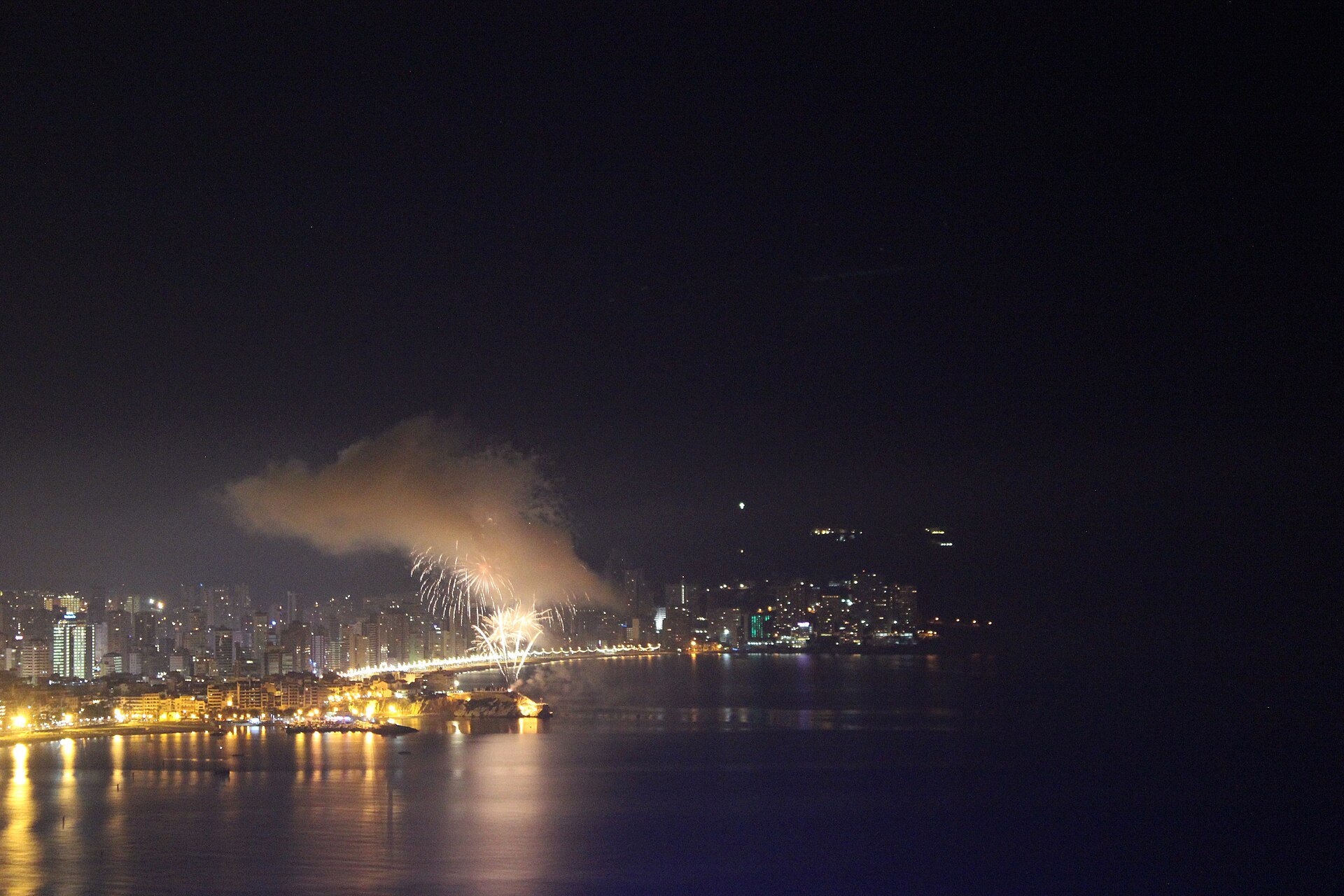 Fireworks bursting over the sea in front of Benidorm's lit seafront at night, seen from across the bay