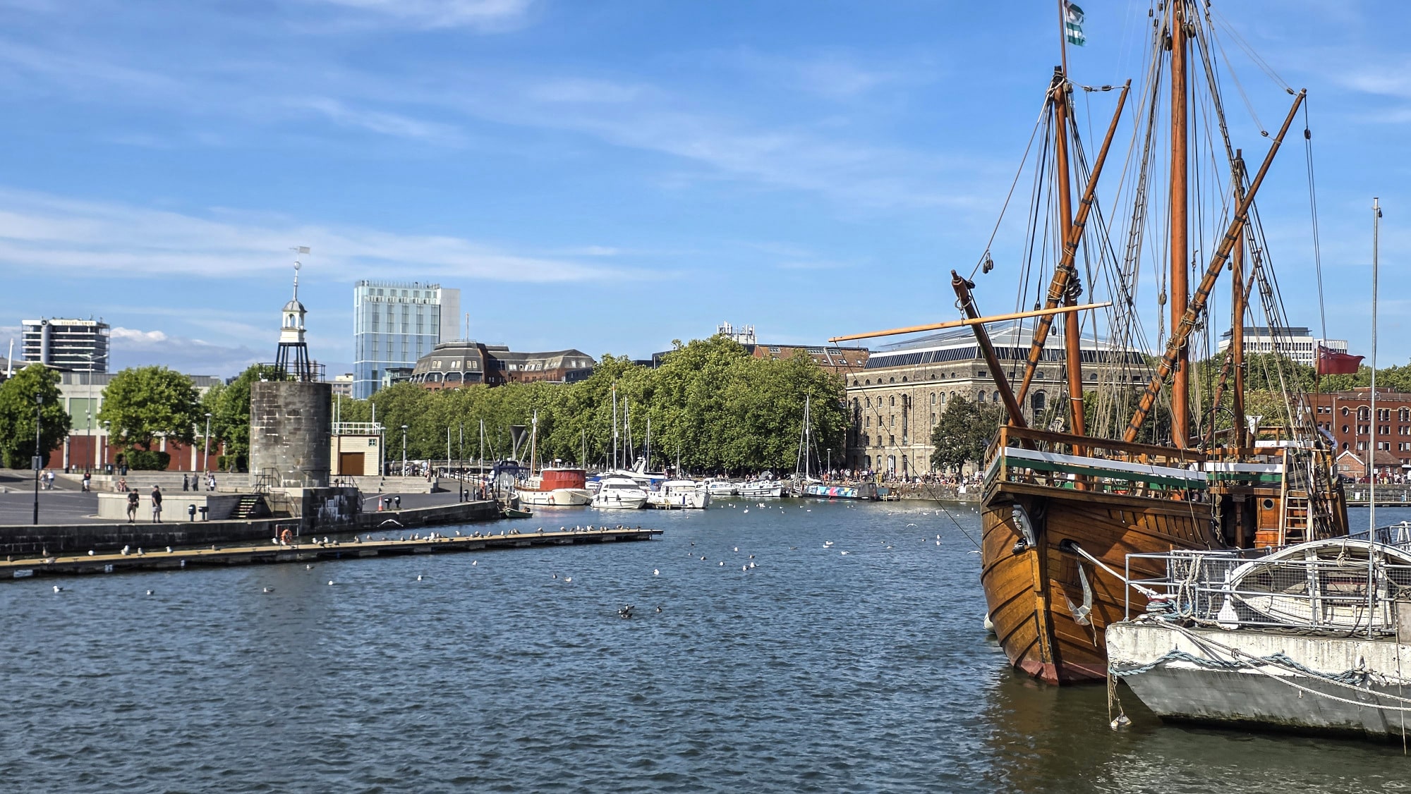 Bristol Harbour, with a wooden sailing ship moored beside a smaller boat, a stone harbour tower and pontoon to the left, and boats along the far quay.