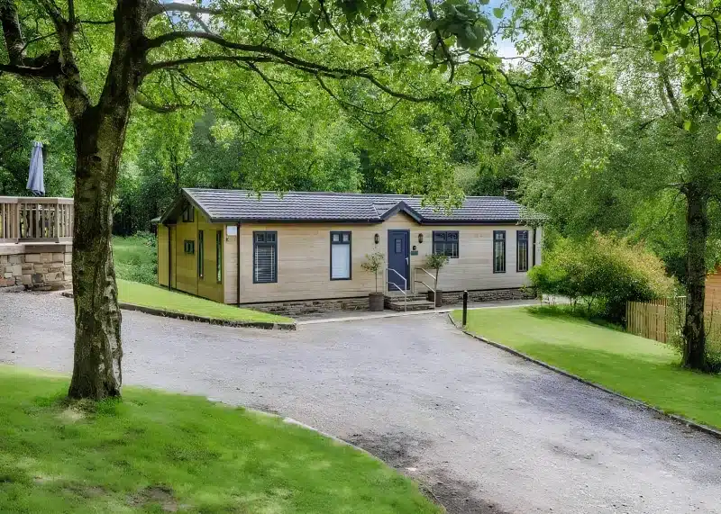 A pale timber lodge with a dark roof and blue door, beside a gravel drive and mature trees.