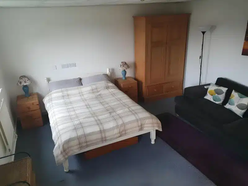 A double bedroom with a plaid-covered bed, pine wardrobe, two bedside lamps and a dark sofa.