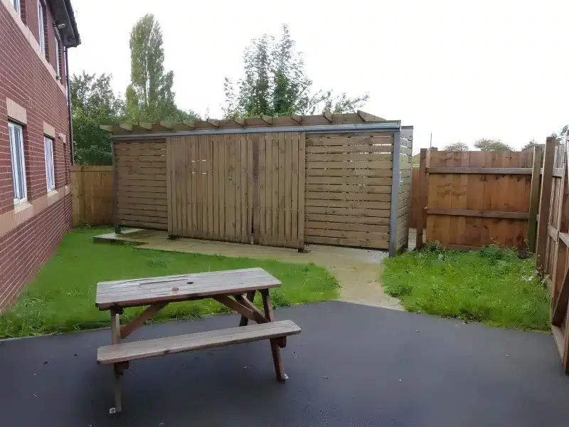 A picnic table on a paved yard beside the brick building, with a wooden screen and grass.