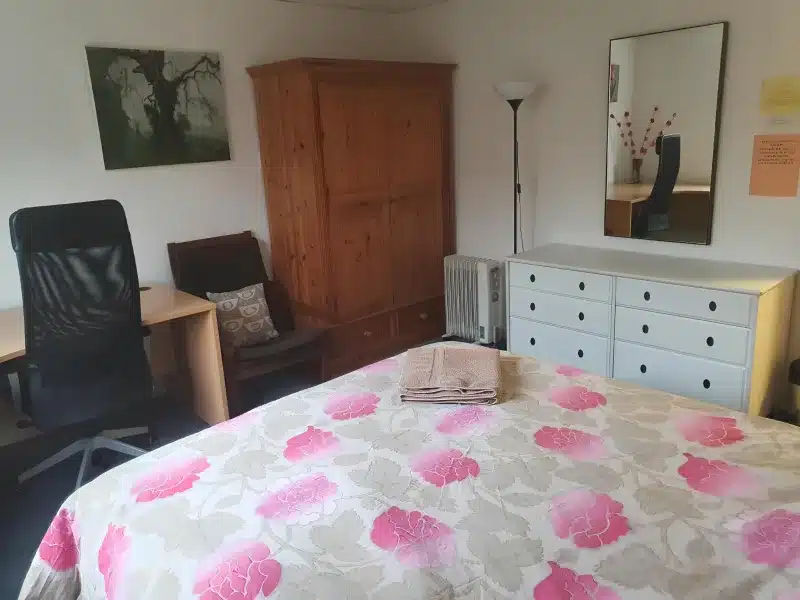 Double room with a pink floral bedspread, a desk with an office chair, an armchair, a pine wardrobe and a white chest of drawers