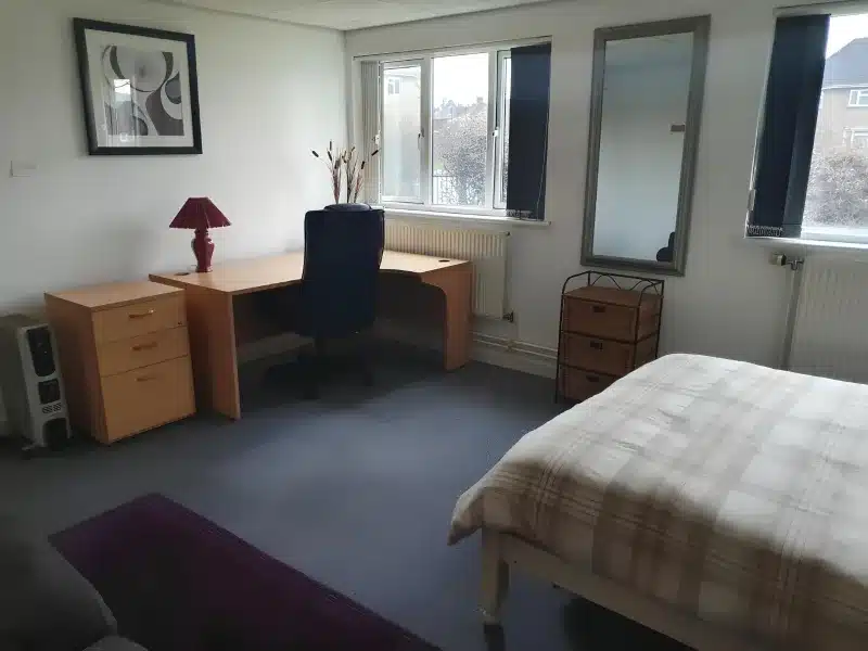 Double room with a corner desk and office chair by two windows, a chest of drawers and a tartan-covered double bed