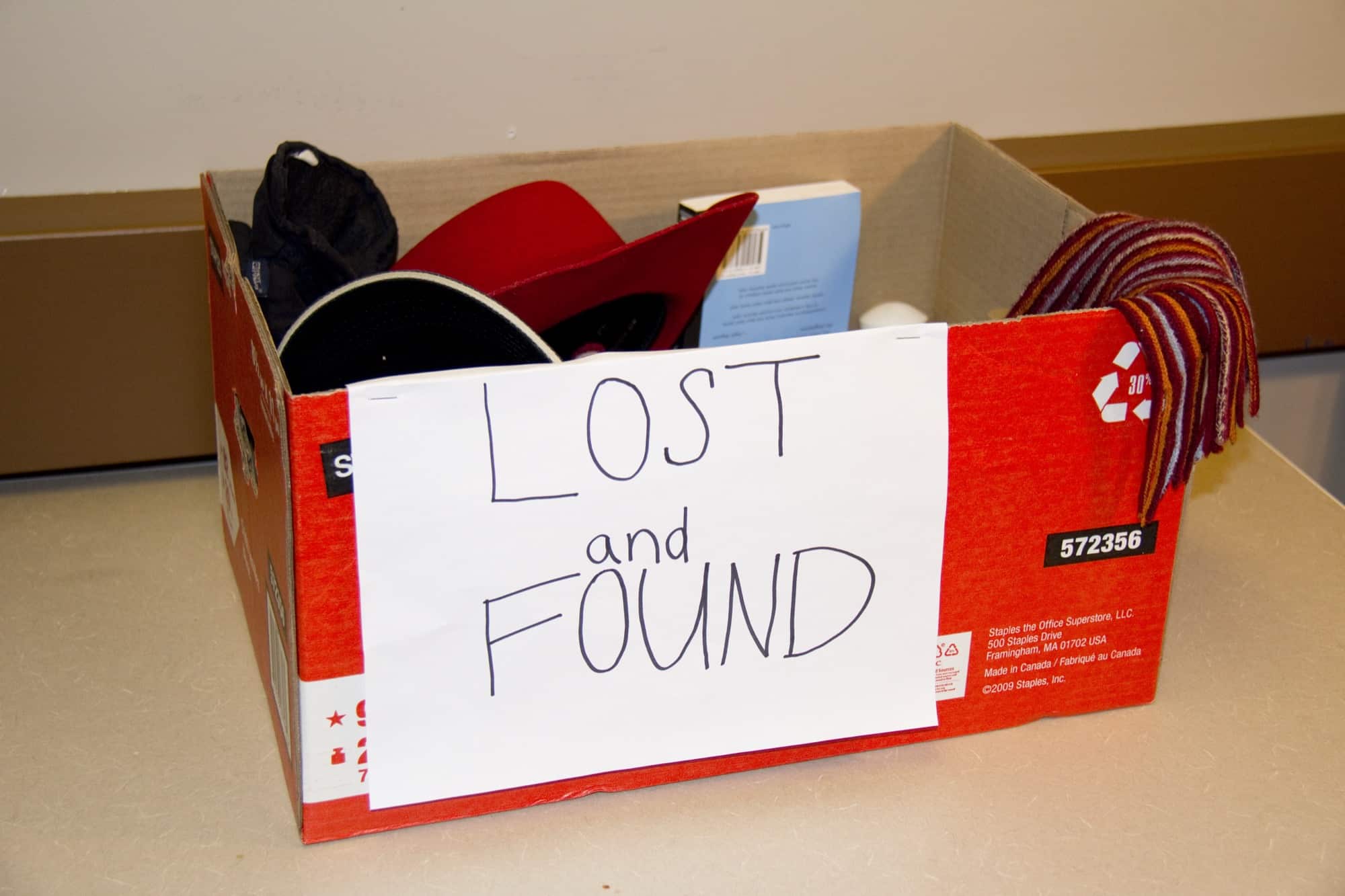 A cardboard box labelled LOST and FOUND in handwriting, holding caps, a book and a scarf