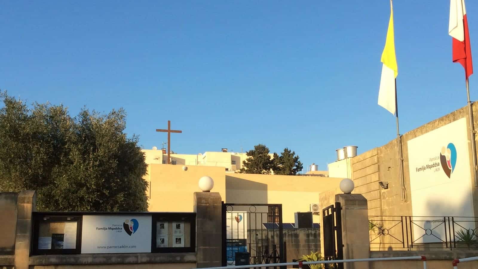 The Iklin parish centre, with the Familja Mqaddsa L-Iklin sign, a metal gate, a cross on the roof, and flags against a clear sky.