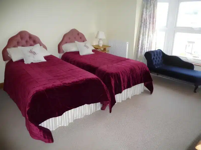 Twin beds with burgundy covers and a blue chaise beside the window