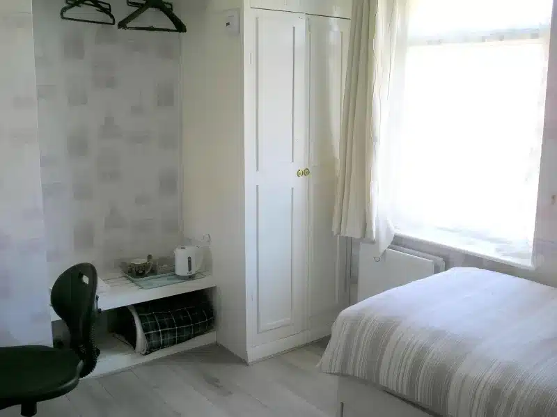 A single bed, white wardrobe, kettle and desk chair in the room at Liverpool Airport Rooms.