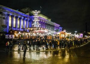Wooden Christmas market stalls and a lit pyramid tower in front of St George's Hall, Liverpool, at night with crowds