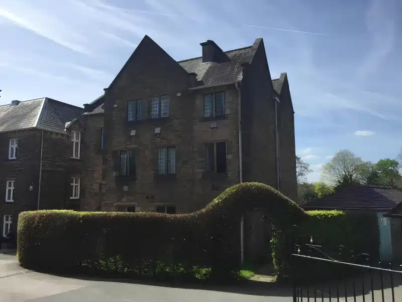 A stone gabled house with leaded windows, a clipped hedge and a black metal gate.