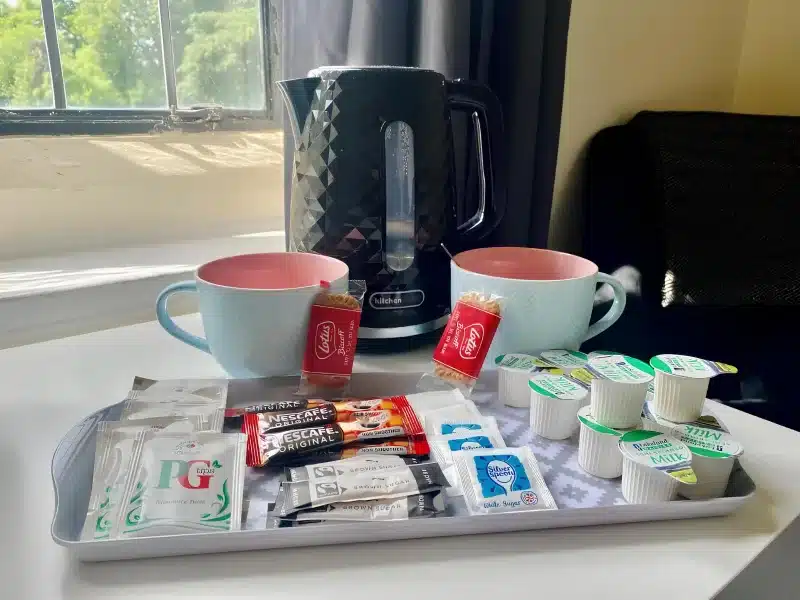 A tray of tea, coffee, sugar and milk pots beside a kettle and two mugs.