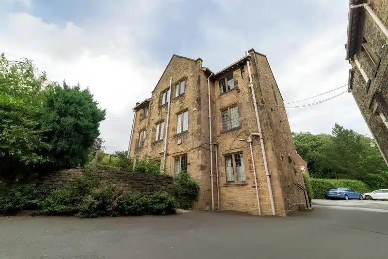 The side of the stone guest house, with a conifer, a dry-stone wall and cars on the drive.