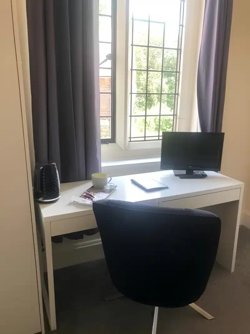 A white desk, a kettle, a monitor and a black chair beside a leaded window.