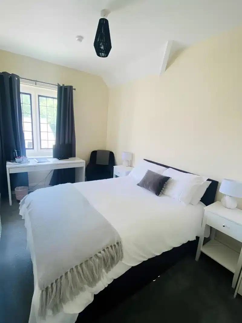 A double bed, a white desk and dark curtains in a cream bedroom.