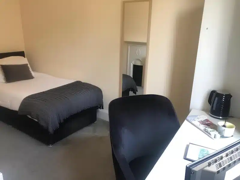 A single bed with a dark throw, a desk, a kettle and a black chair.