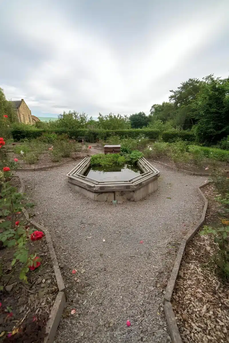 Gravel paths around a small formal pond, with roses and the stone house beyond.