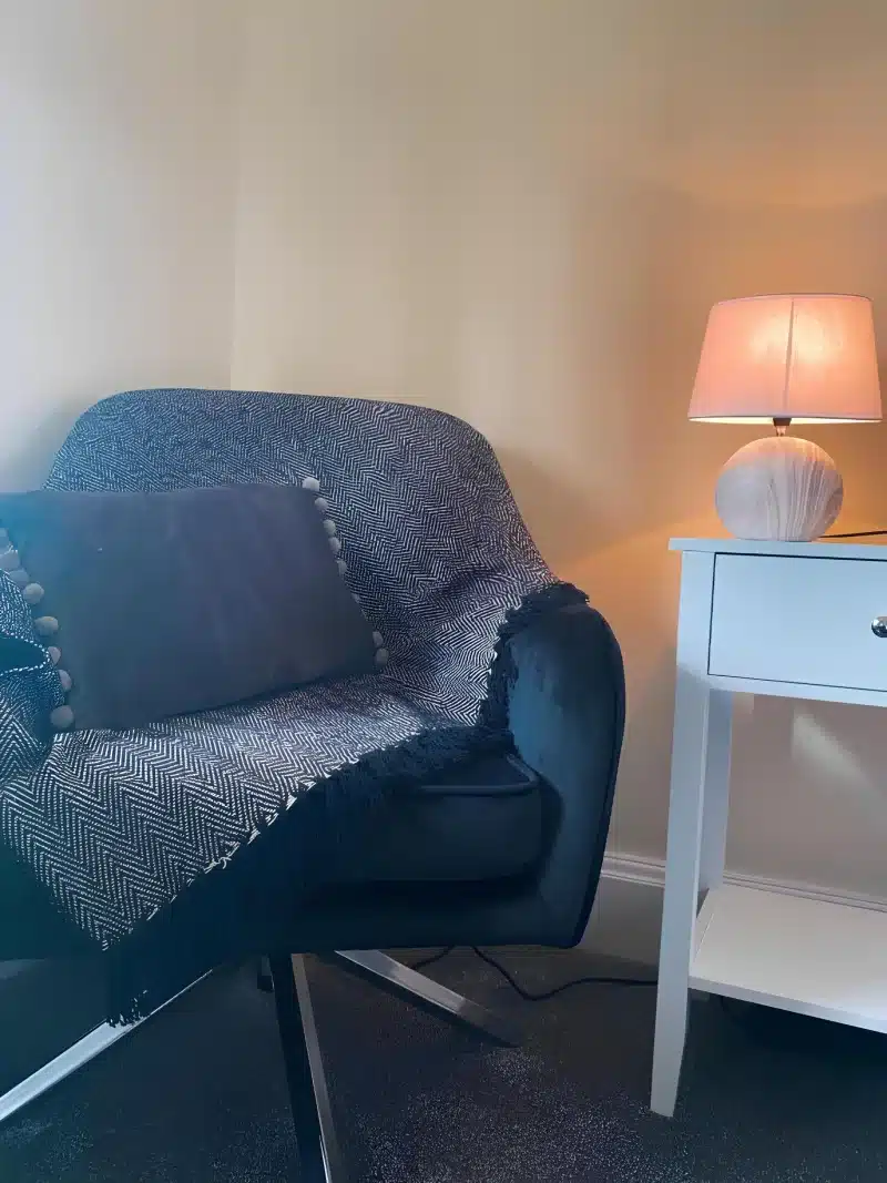 A dark armchair with a herringbone throw beside a white table and a lamp.