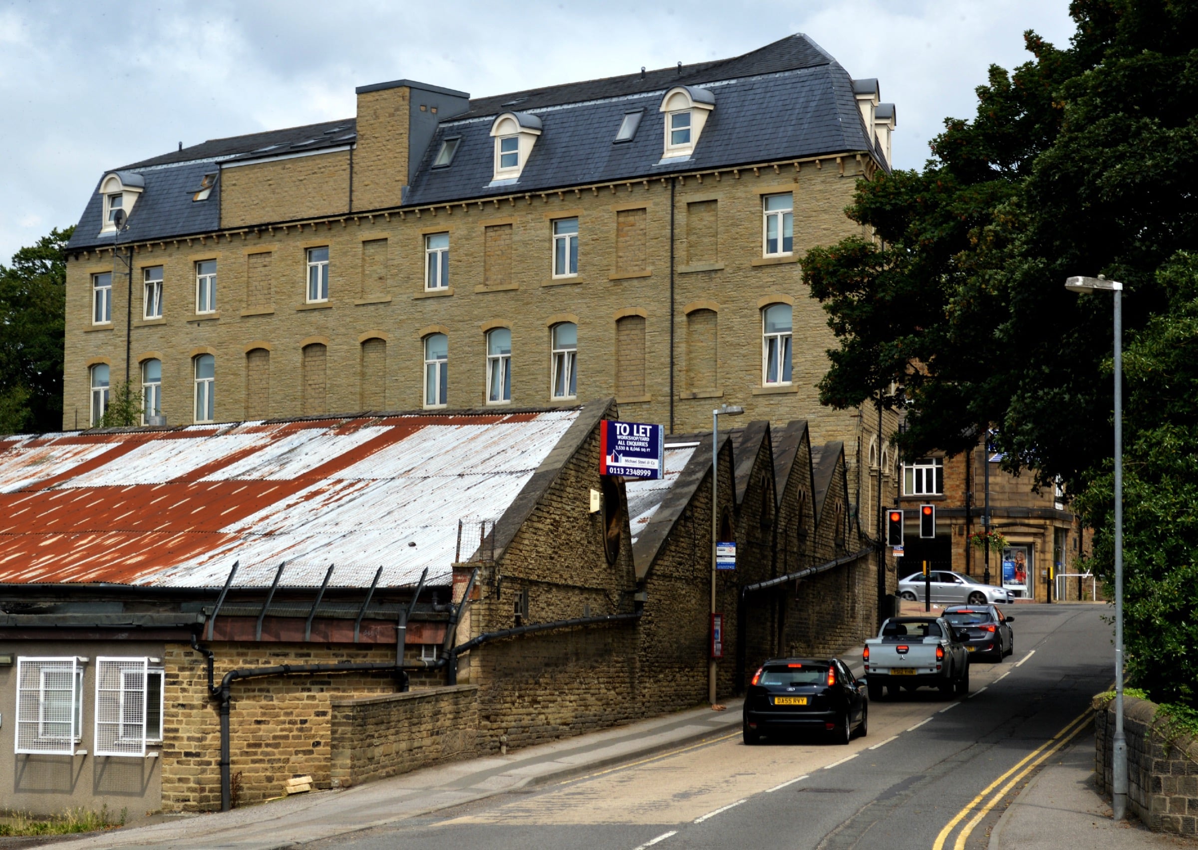 A tall sandstone mill with a dark slate roof stands above lower workshops with a rusted corrugated roof, beside a two-lane road of queued cars in Mirfield.