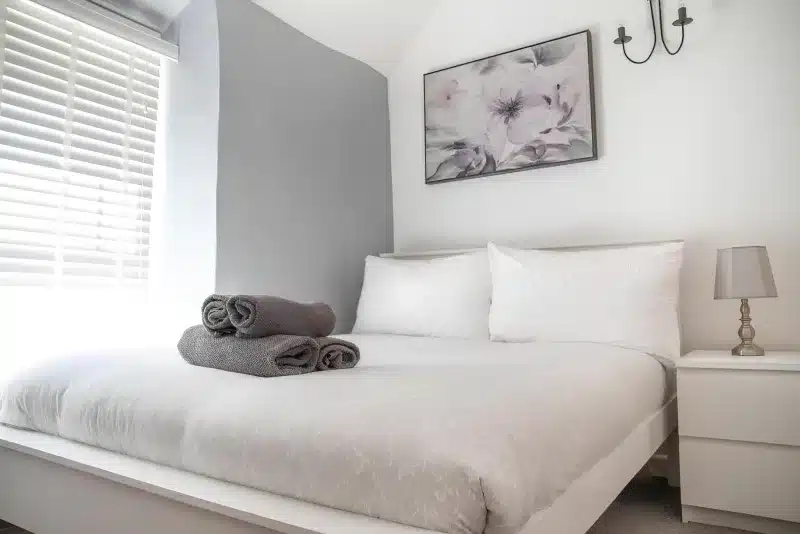 A white double bed with grey towels, a bedside lamp and a floral picture above the pillows.
