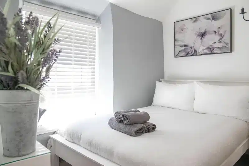 A white double bed with grey towels folded on it, a window with white blinds, and a floral picture.