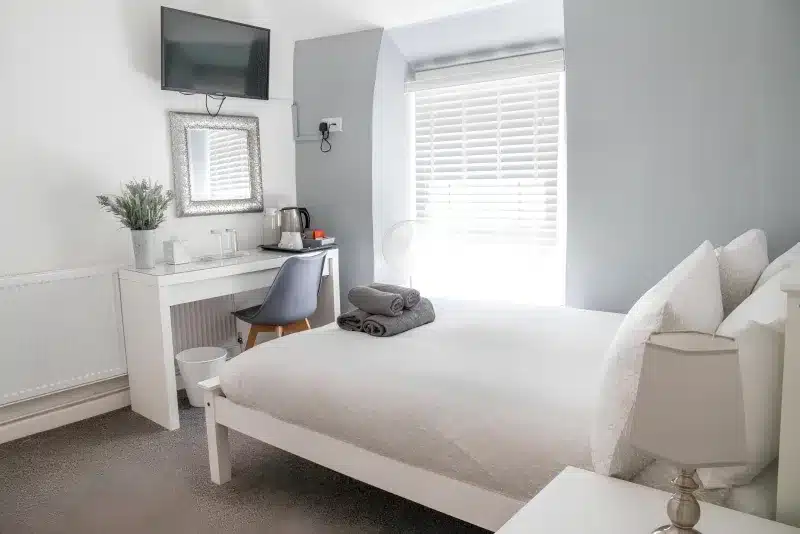 A white double bed, a desk with a kettle, a television and a bright window.
