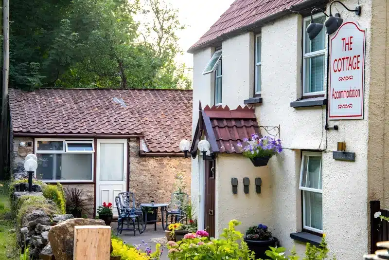 Outdoor chairs and a table in front of The Cottage, beside a low stone wall and the accommodation sign.