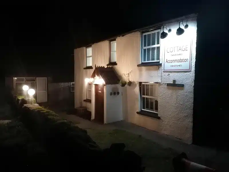 The Cottage at night from the side approach, with the sign lit and a dark hedge in front.