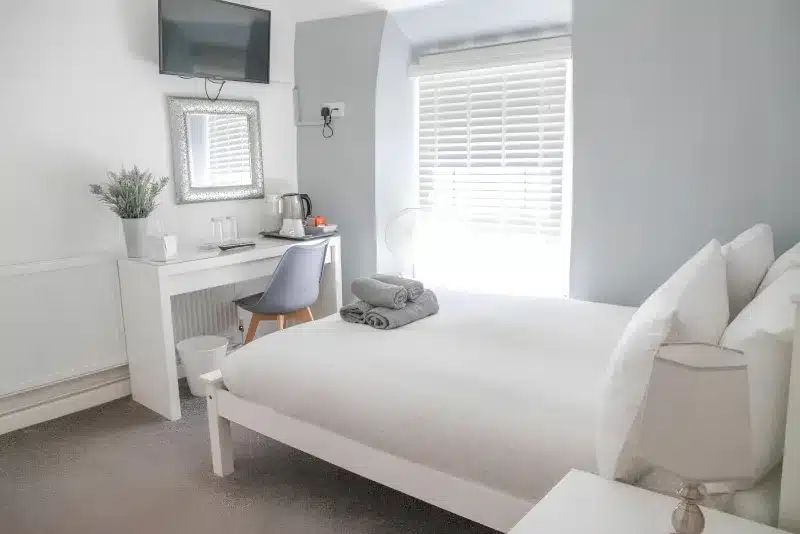 A white double bed beside a desk with a kettle, a wall-mounted television and a bright window.