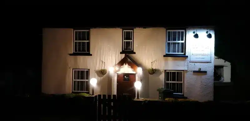 The Cottage at night, with the accommodation sign lit and round lamps by the front door.