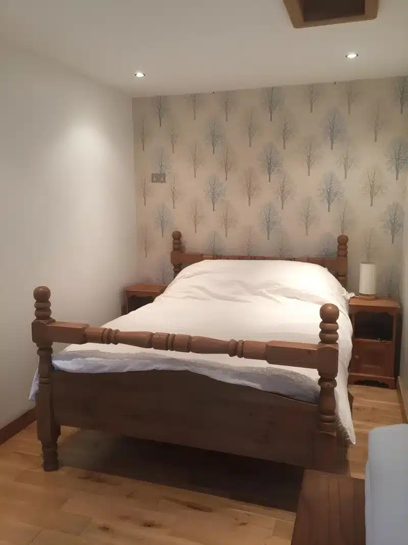 Close view of the wooden bed, white bedding and tree wallpaper.