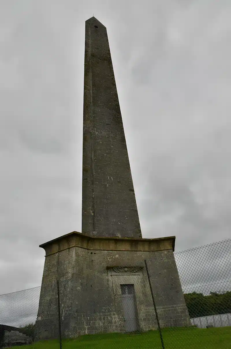 The stone Wellington Monument against a grey sky, with a fence across the grass at its base.